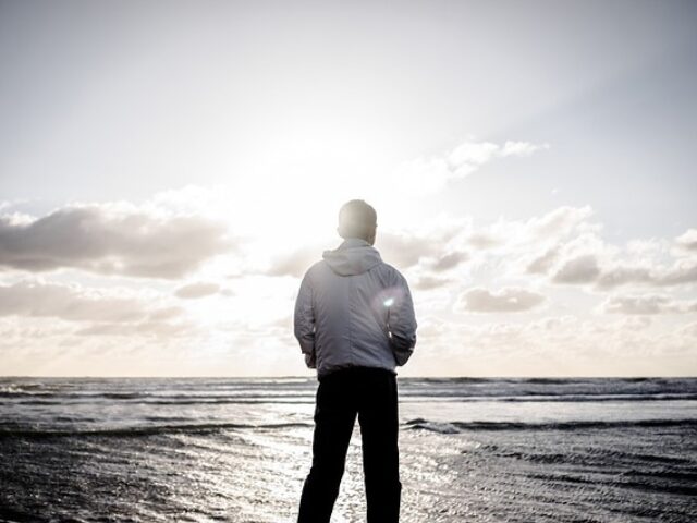 海辺で立ち止まり、遠くの水平線を見つめる男性の後ろ姿。彼は白いジャケットを着ており、日差しが彼の背後から差し込んでいる。空には雲が広がり、穏やかな海が広がる風景が広がっている画像